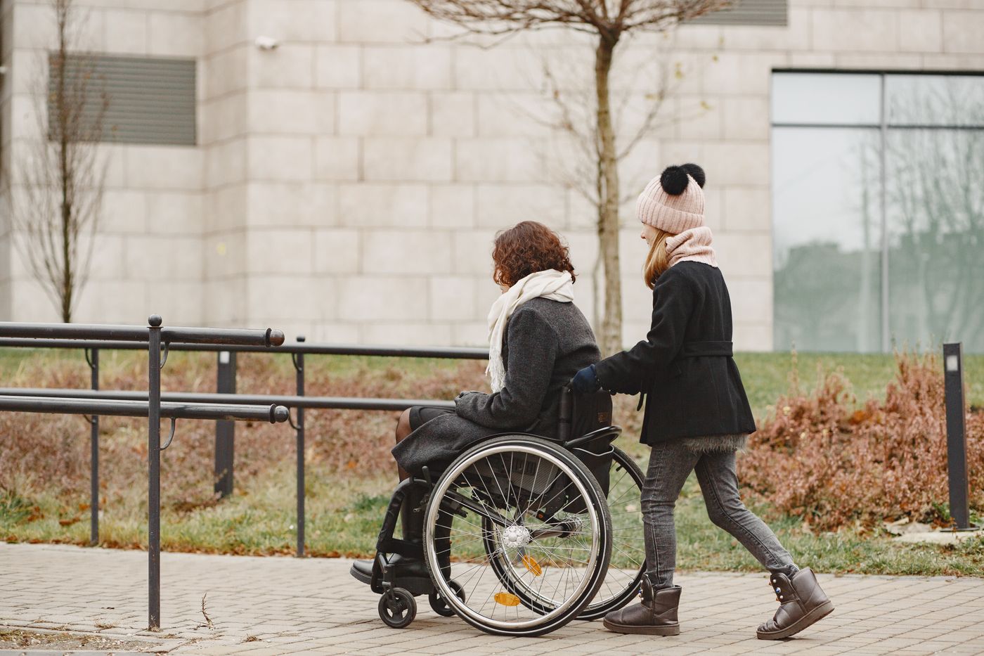 A passenger in a wheelchair accompanied along an accessible path toward a building entrance on an overcast day.